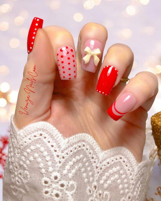 Ensemble de press-on nails rouges et nude décorés de pois blancs, de french rouges et d’un nœud 3D blanc, présenté sur une main féminine avec un fond lumineux et festif
