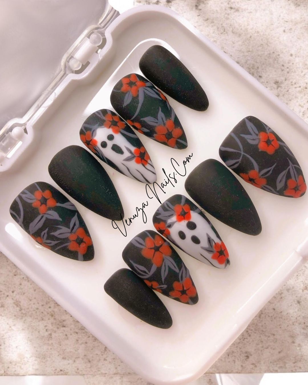 photo avec flash, dans sa boite blanche, le set de press on nails est noir mat avec un feuillage gris orné de fleurs oranges avec sur le majeur un fantôme hurlant.