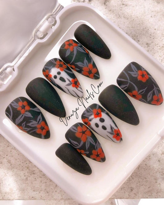 photo avec flash, dans sa boite blanche, le set de press on nails est noir mat avec un feuillage gris orné de fleurs oranges avec sur le majeur un fantôme hurlant.
