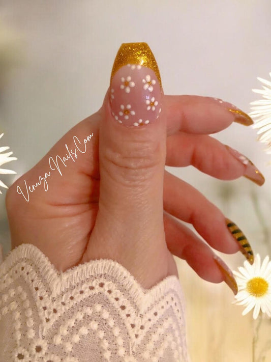 Manucure press-on rose et or avec détails floraux et abeille scintillante, portée sur main, rendu chic et lumineux. Venuza Nails