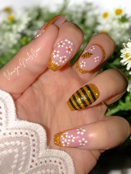 Manucure press-on rose et or avec détails floraux et abeille scintillante, portée sur main, rendu chic et lumineux. Venuza Nails