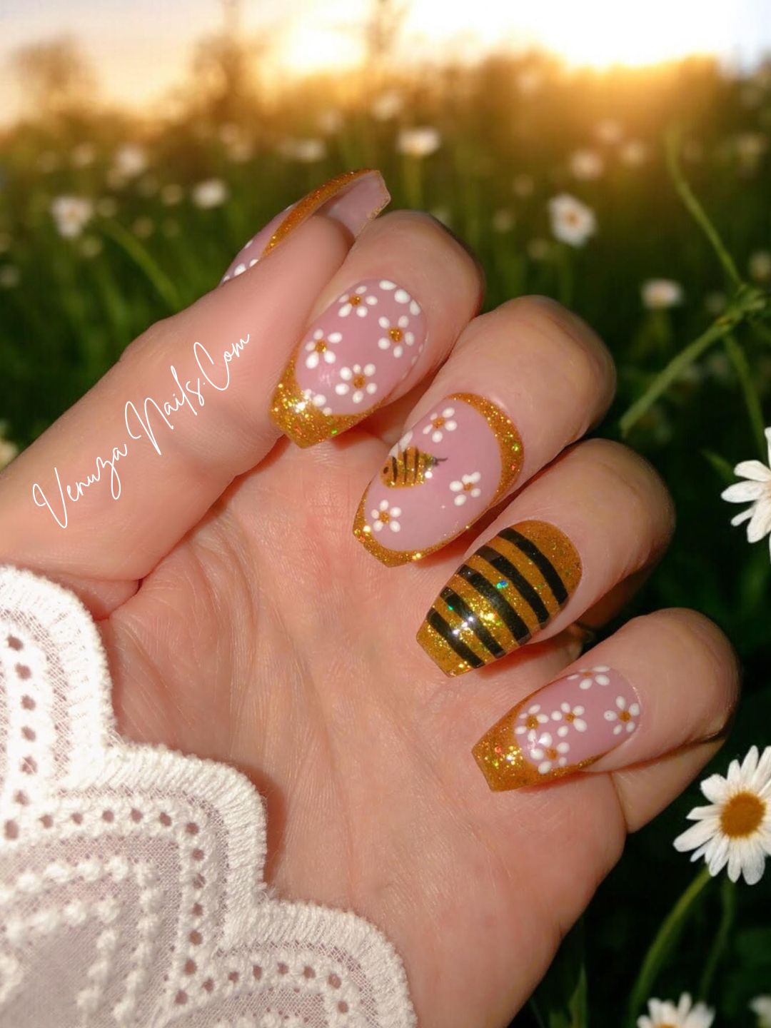 Manucure press-on rose et or avec détails floraux et abeille scintillante, portée sur main, rendu chic et lumineux. Venuza Nails