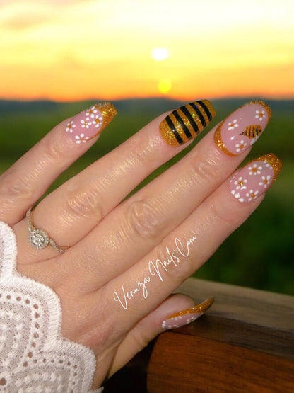 Manucure press-on rose et or avec détails floraux et abeille scintillante, portée sur main, rendu chic et lumineux. Venuza Nails