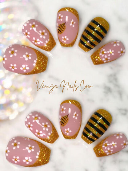 Press-on nails rose poudré avec french pailletée dorée, fleurs blanches et motif abeille doré, présentés à plat sur fond marbre – Venuza Nails.