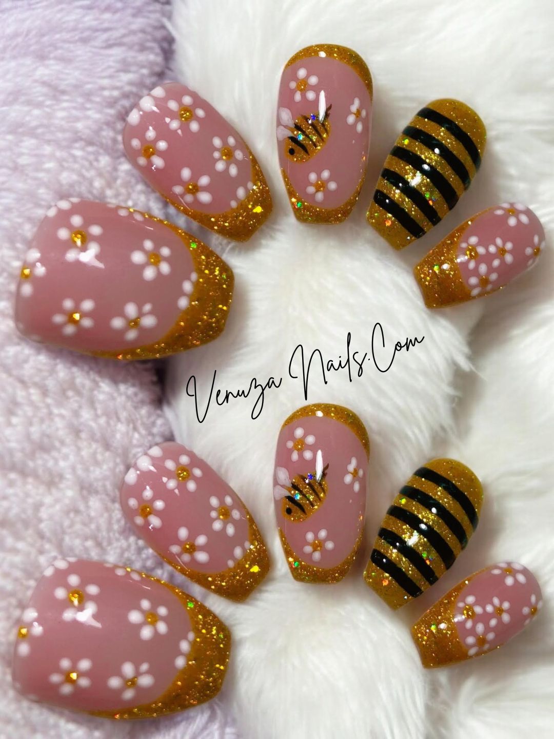 Press-on nails rose poudré avec french pailletée dorée, fleurs blanches et motif abeille doré, présentés à plat sur fond couverture moelleuse – Venuza Nails.