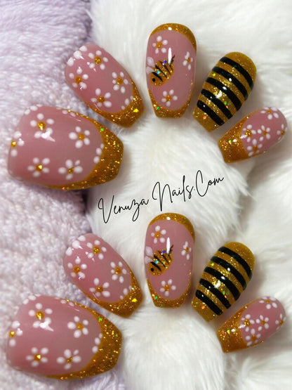 Press-on nails rose poudré avec french pailletée dorée, fleurs blanches et motif abeille doré, présentés à plat sur fond couverture moelleuse – Venuza Nails.