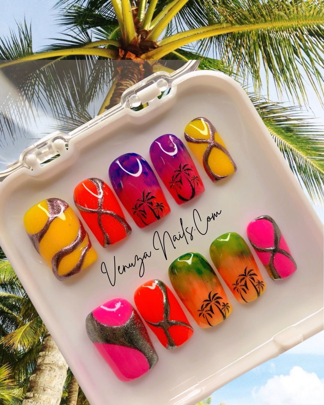 Press-on nails Caribbean Vibes aux dégradés tropicaux rose, violet et orange avec motifs palmiers noirs et détails métallisées argentées, fond coucher de soleil violet et palmiers.