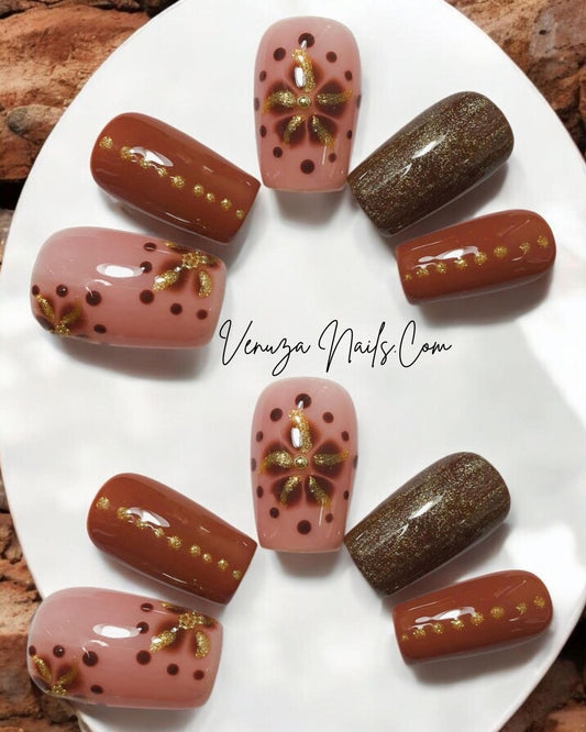 Ensemble de faux ongles press-on marron, rose poudré et doré avec motifs floraux scintillants, disposés sur un fond marroné et blanc.