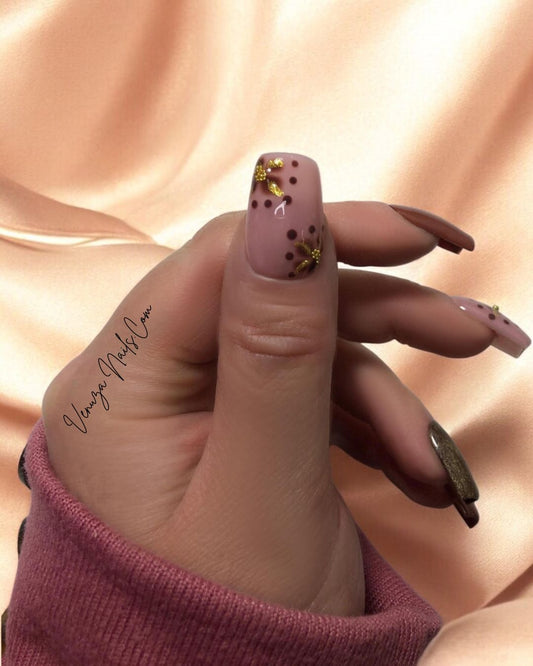 Main portant des faux ongles press-on marron, rose poudré et doré avec motifs floraux dorés scintillants, prise sur un fond marroné et blanc.