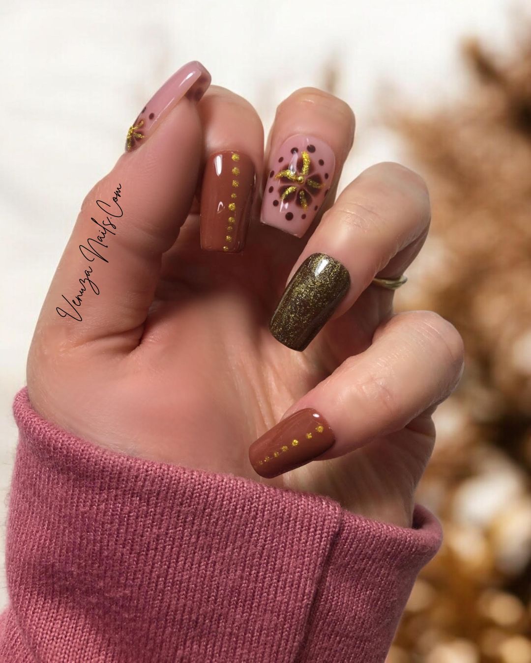 Main portant des faux ongles press-on marron, rose poudré et doré avec motifs floraux dorés scintillants, prise sur un fond marroné et blanc.