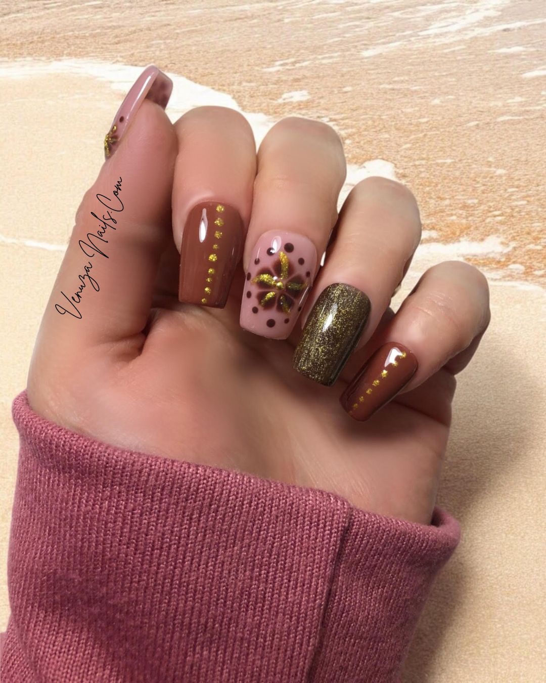 Main portant des faux ongles press-on marron, rose poudré et doré avec motifs floraux dorés scintillants, prise sur un fond marroné et blanc.