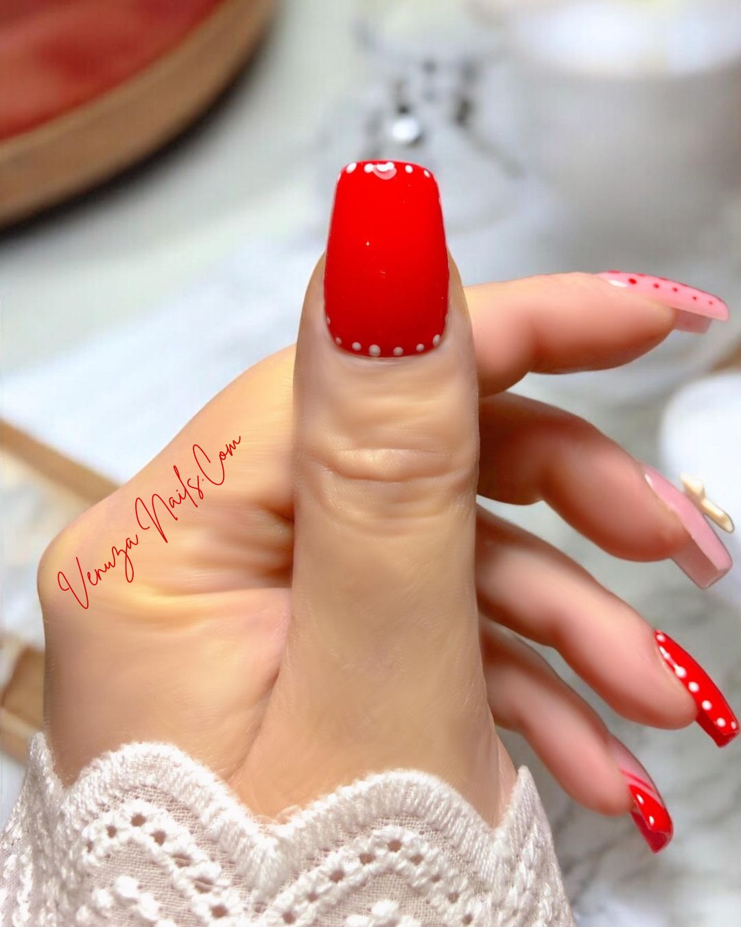 Ensemble de press-on nails rouges et nude décorés de pois blancs, de french rouges et d’un nœud 3D blanc, présenté sur une main féminine avec un fond lumineux et festif