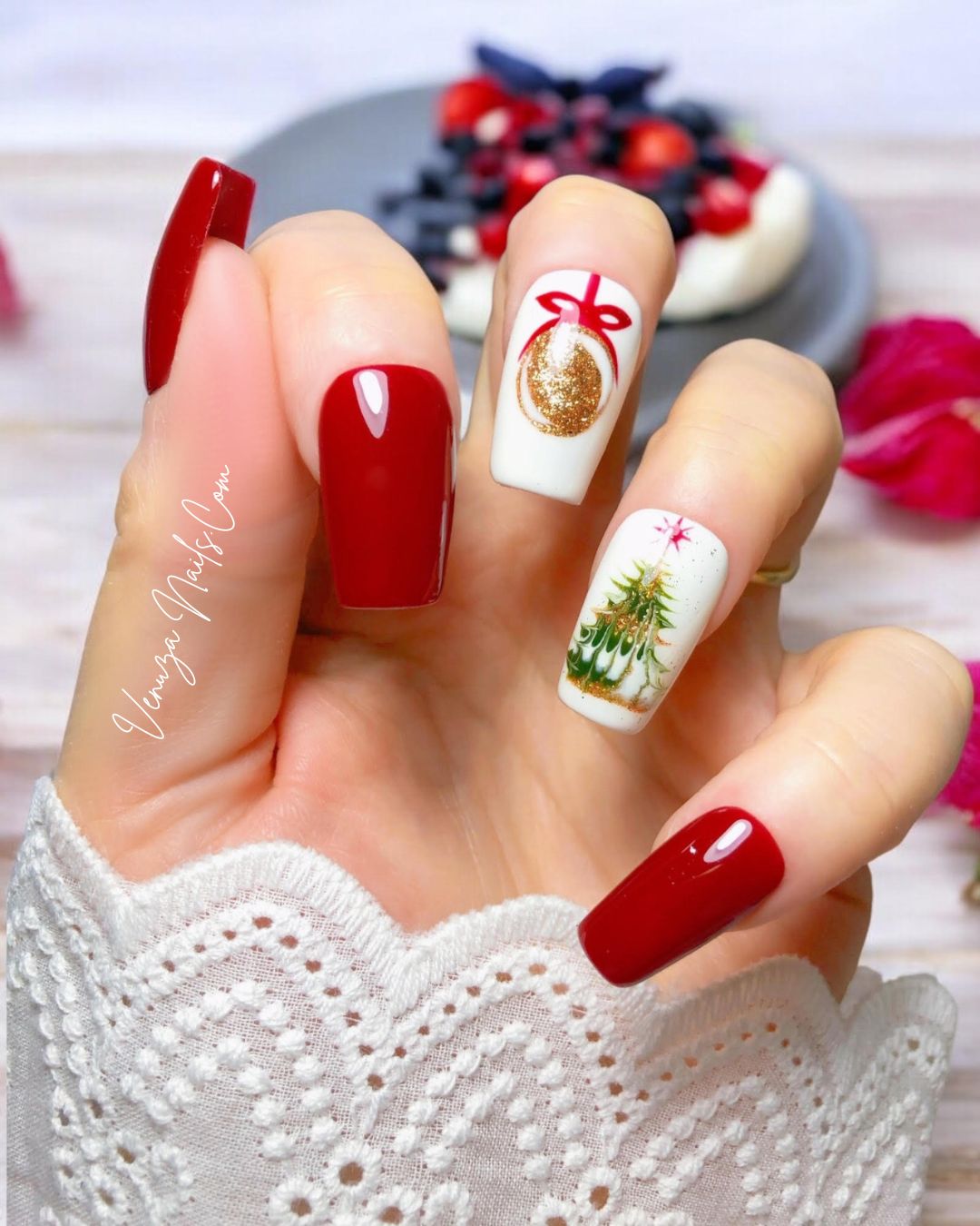 Main portant des press-on nails rouges et blancs au style Noël. Deux ongles sont rouges brillants, un ongle blanc avec une boule dorée dessinée avec un ruban rouge, et un autre ongle blanc décoré d’un petit sapin de Noël doré et vert. Les capsules sont longues et brillantes, avec une ambiance festive et élégante.
