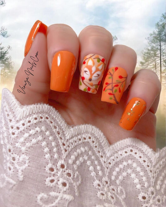 Modèle « Rêve d’Automne » de Venuza Nails porté sur une main féminine : press-on nails orange élégants ornés d’éclats de feuilles d’or, d’un motif de renarde endormie et de branches garnies de baies, pour une manucure automnale poétique et raffinée.