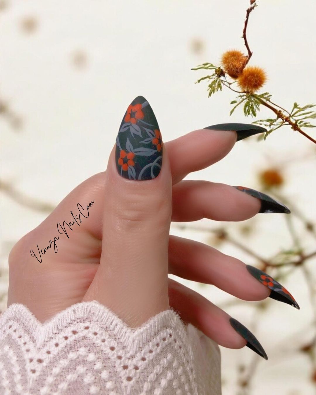 main de femme portant un set de press on nails d'halloween sur fond de fleurs orangées, le set est noir mat avec un feuillage gris orné de fleurs oranges avec sur le majeur un fantôme hurlant.