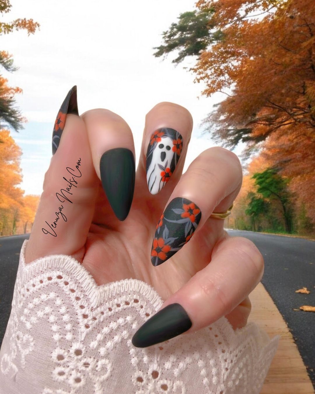 main de femme portant un set de press on nails d'halloween sur fond de feuilles de sapin fanées, le set est noir mat avec un feuillage gris orné de fleurs oranges avec sur le majeur un fantôme hurlant.