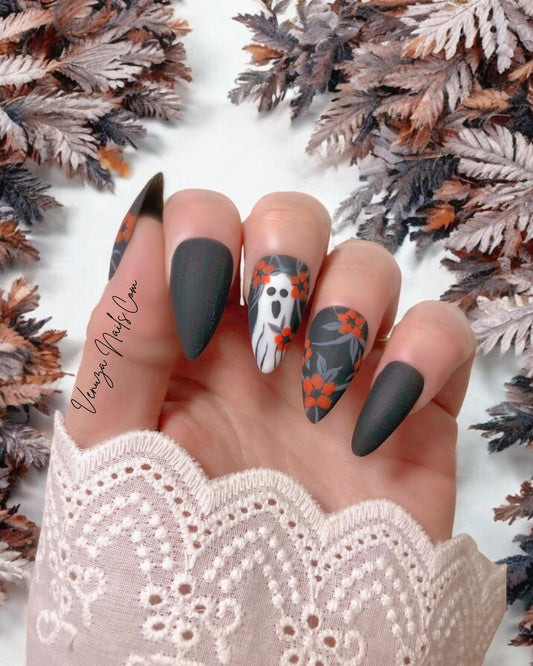 main de femme portant un set de press on nails d'halloween sur fond de feuilles de sapin fanées, le set est noir mat avec un feuillage gris orné de fleurs oranges avec sur le majeur un fantôme hurlant.