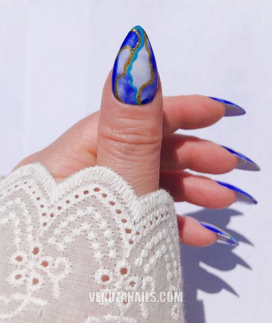 (main avec ongles marbrés bleu, blanc et or) :
alt="Press-on nails marbrés bleu, blanc et or – faux ongles en forme amande de la marque Venuza Nails"