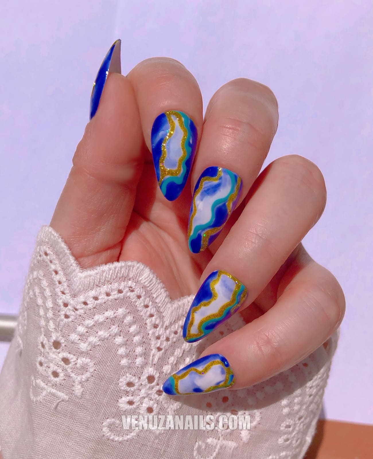 (main avec ongles marbrés bleu, blanc et or) :
alt="Press-on nails marbrés bleu, blanc et or – faux ongles en forme amande de la marque Venuza Nails"