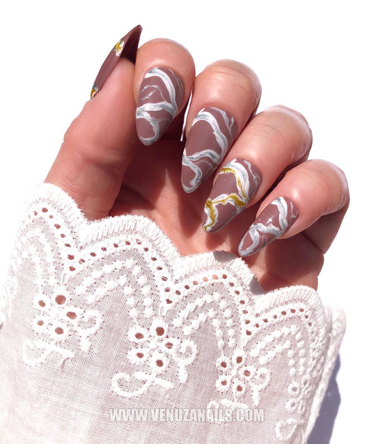 Main portant des faux ongles press-on de la collection Venuza Nails, design marbré couleur marron avec détails dorés et blancs, finition brillante, forme amande – modèle "Luxor".