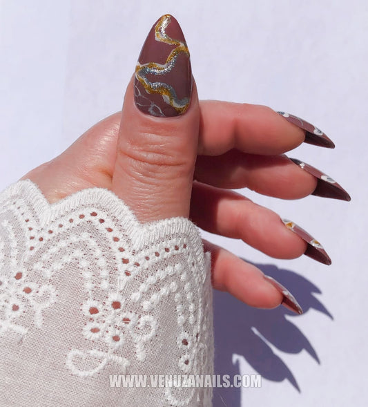 Main portant des faux ongles press-on de la collection Venuza Nails, design marbré couleur marron avec détails dorés et blancs, finition brillante, forme amande – modèle "Luxor".