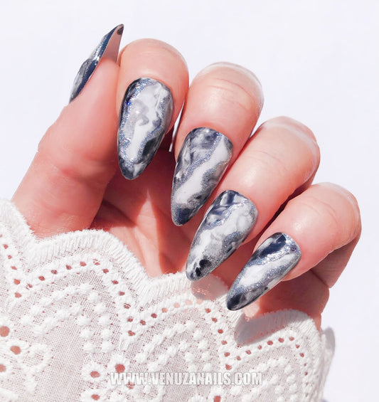 Mains portant des press-on nails marbrés noirs et argentés “Mystic Black”, finition brillante avec des détails métallisés, design élégant et sophistiqué, idéal pour un look chic et audacieux.