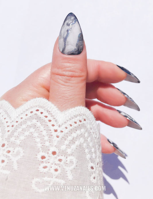 Mains portant des press-on nails marbrés noirs et argentés “Mystic Black”, finition brillante avec des détails métallisés, design élégant et sophistiqué, idéal pour un look chic et audacieux.