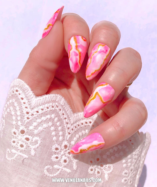 main montrant les faux ongles faits main, marbrés, rose, or, blanc, avec petite manchette en dentelle blanche.