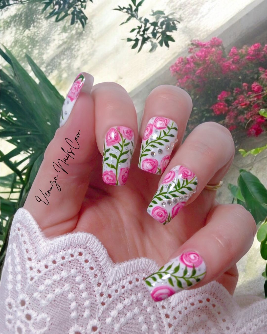 Press-on nails décorés de roses roses peintes à la main, feuillages verts et strass brillants sur fond blanc, design floral élégant et romantique.