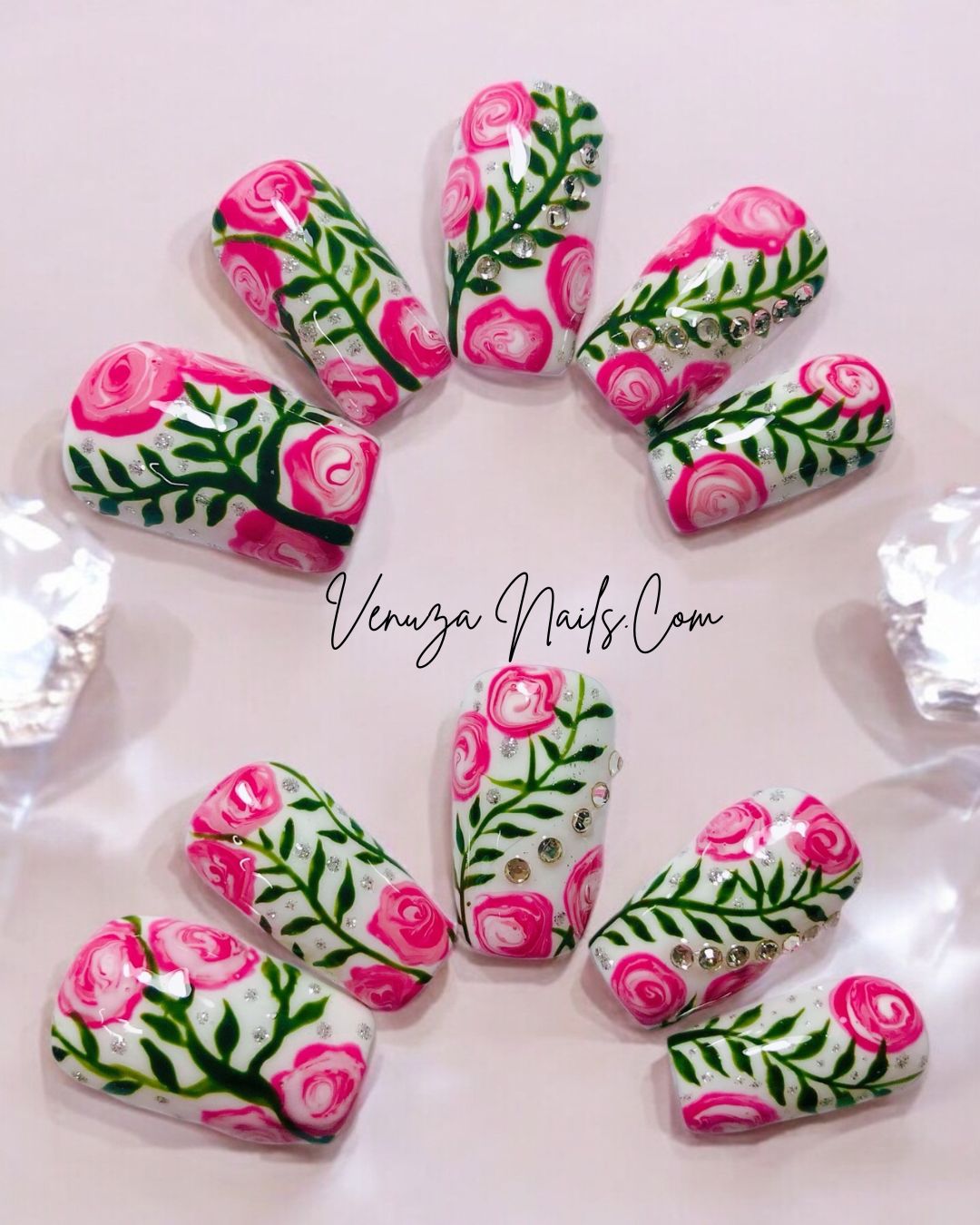 Press-on nails décorés de roses roses peintes à la main, feuillages verts et strass brillants sur fond blanc, design floral élégant et romantique.
