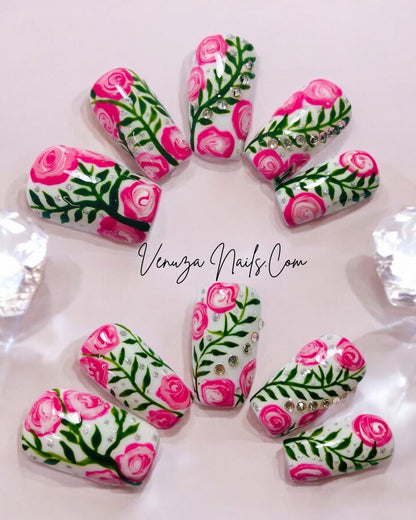 Press-on nails décorés de roses roses peintes à la main, feuillages verts et strass brillants sur fond blanc, design floral élégant et romantique.