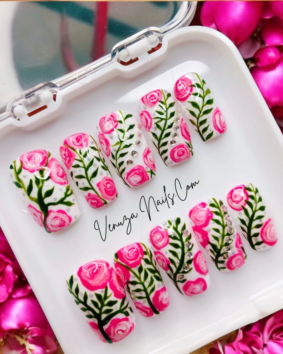 Press-on nails décorés de roses roses peintes à la main, feuillages verts et strass brillants sur fond blanc, design floral élégant et romantique.
