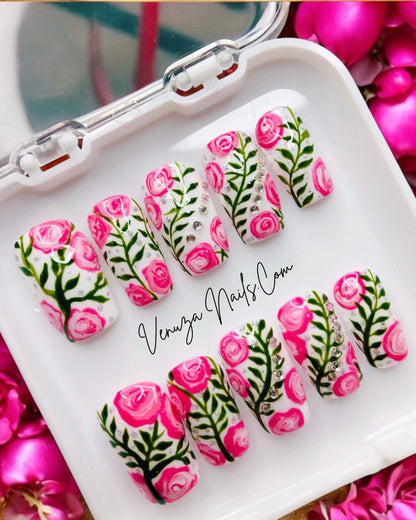 Press-on nails décorés de roses roses peintes à la main, feuillages verts et strass brillants sur fond blanc, design floral élégant et romantique.