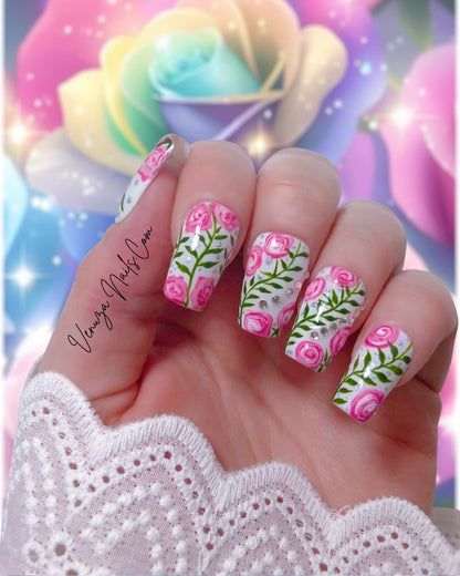 Press-on nails décorés de roses roses peintes à la main, feuillages verts et strass brillants sur fond blanc, design floral élégant et romantique.