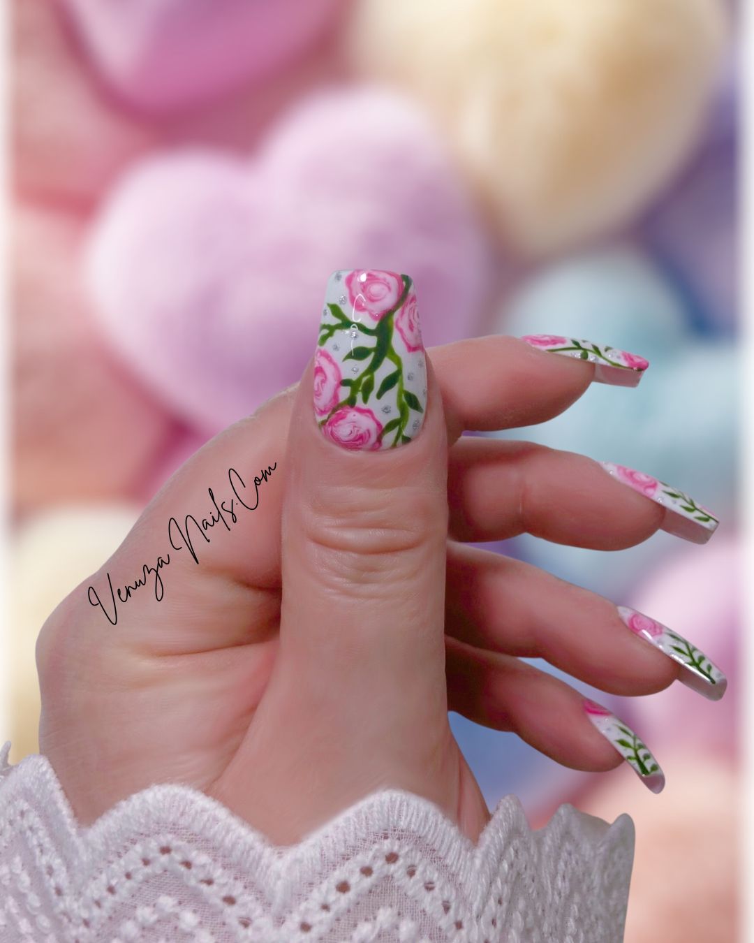 Press-on nails décorés de roses roses peintes à la main, feuillages verts et strass brillants sur fond blanc, design floral élégant et romantique.