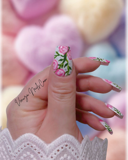 Press-on nails décorés de roses roses peintes à la main, feuillages verts et strass brillants sur fond blanc, design floral élégant et romantique.
