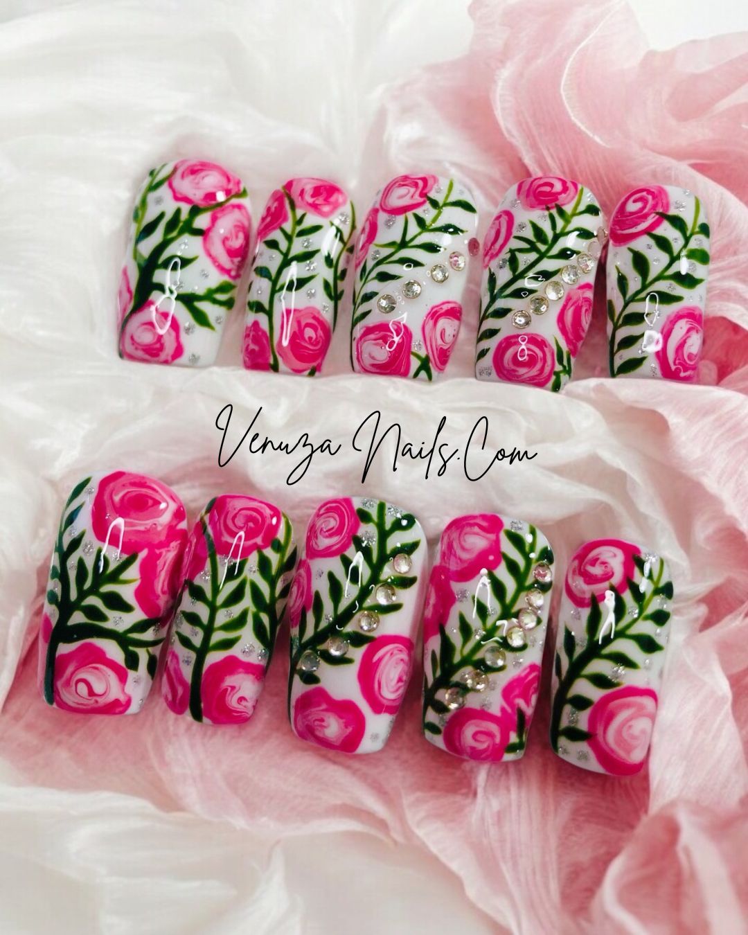 Press-on nails décorés de roses roses peintes à la main, feuillages verts et strass brillants sur fond blanc, design floral élégant et romantique.