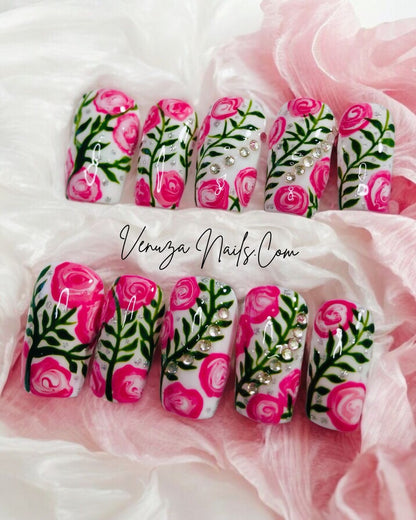 Press-on nails décorés de roses roses peintes à la main, feuillages verts et strass brillants sur fond blanc, design floral élégant et romantique.